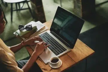 A person typing on a laptop at a wooden table, surrounded by books, sunglasses, and a cup of coffee in a cozy café setting.
