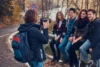 A person with a backpack photographs four smiling friends sitting on a roadside railing in a forested autumn landscape, creating a cheerful and relaxed atmosphere.