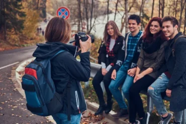 A person with a backpack photographs four smiling friends sitting on a roadside railing in a forested autumn landscape, creating a cheerful and relaxed atmosphere.