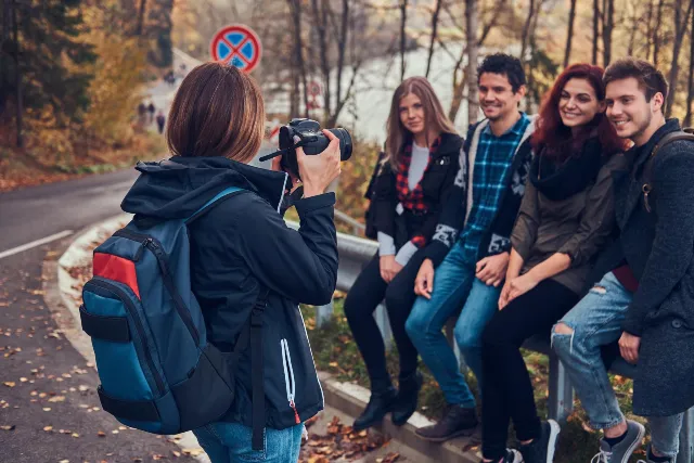 A person with a backpack photographs four smiling friends sitting on a roadside railing in a forested autumn landscape, creating a cheerful and relaxed atmosphere.