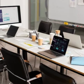 A modern conference room with a long table cluttered with laptops, charts, papers, and coffee cups. The setting is busy and professional, suggesting a work meeting.
