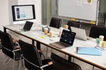 A modern conference room with a long table cluttered with laptops, charts, papers, and coffee cups. The setting is busy and professional, suggesting a work meeting.