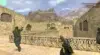 Video game screenshot from a desert map in Counter-Strike. Players aim guns in a tense combat scene with stone walls and mountain backdrop.