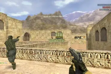 Video game screenshot from a desert map in Counter-Strike. Players aim guns in a tense combat scene with stone walls and mountain backdrop.