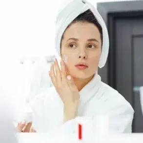 A woman wearing a bathrobe and towel turban applies face cream, looking at herself in a bathroom mirror. The scene conveys a calm, self-care routine.