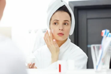 A woman wearing a bathrobe and towel turban applies face cream, looking at herself in a bathroom mirror. The scene conveys a calm, self-care routine.