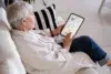 An elderly person sits on a white couch, using a tablet with a bright interface. They appear focused and relaxed, with a striped cushion beside them.