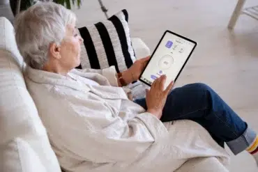 An elderly person sits on a white couch, using a tablet with a bright interface. They appear focused and relaxed, with a striped cushion beside them.