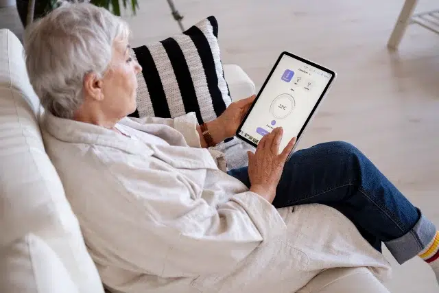 An elderly person sits on a white couch, using a tablet with a bright interface. They appear focused and relaxed, with a striped cushion beside them.