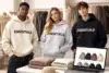 Three models wearing "ESSENTIALS" hoodies in beige, grey, and black, surrounded by clothes and a laptop displaying similar apparel. Casual, trendy vibe.