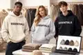 Three models wearing "ESSENTIALS" hoodies in beige, grey, and black, surrounded by clothes and a laptop displaying similar apparel. Casual, trendy vibe.