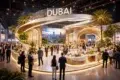 Spacious expo hall with a glowing "Dubai" sign, featuring a futuristic design, lush greenery, and a cityscape backdrop. Attendees in business attire network.