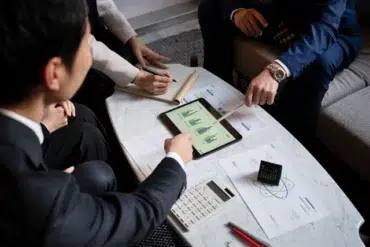 A group of professionals in suits collaborates over a tablet displaying graphs, surrounded by documents and a calculator on a table.