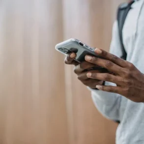 A person in a gray hoodie uses a smartphone outdoors against a blurred wood-like wall. The scene conveys a relaxed and focused mood.