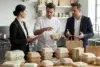Three people discuss eco-friendly packaging in a kitchen. A woman holds a tablet, a chef gestures to boxes, and a man in a suit listens. Mood is collaborative.