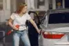 A woman in a white shirt and jeans refuels a silver car at a gas station. She holds a red pump nozzle, with the car door open, appearing focused.
