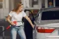 A woman in a white shirt and jeans refuels a silver car at a gas station. She holds a red pump nozzle, with the car door open, appearing focused.