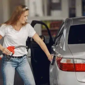 A woman in a white shirt and jeans refuels a silver car at a gas station. She holds a red pump nozzle, with the car door open, appearing focused.