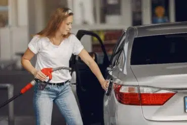 A woman in a white shirt and jeans refuels a silver car at a gas station. She holds a red pump nozzle, with the car door open, appearing focused.