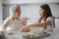 An elderly woman and a young woman share a light moment in a bright kitchen. The older woman gestures animatedly while the young woman eats a cookie, smiling.