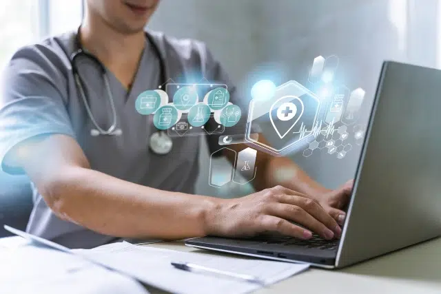 A healthcare professional in scrubs types on a laptop with futuristic, glowing medical icons above the keyboard, conveying innovation and technology.