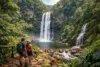 A couple stands admiring a stunning waterfall surrounded by thick green jungle and rocky terrain, with other hikers in the background.