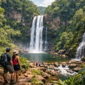 A couple stands admiring a stunning waterfall surrounded by thick green jungle and rocky terrain, with other hikers in the background.