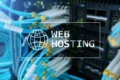 Blurry close-up of blue network cables connected to a server. Overlay text reads "Web Hosting" with a globe icon, conveying a tech-focused theme.