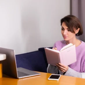 A woman in a lavender sweater sits at a table, attentively reading a pink notebook. Beside her are a silver laptop and smartphone, suggesting a calm, focused work or study session.