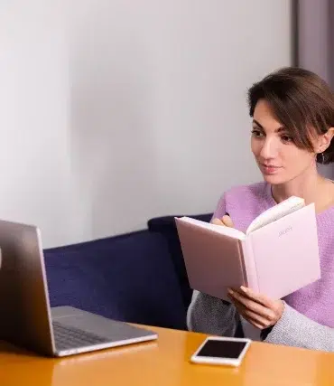 A woman in a lavender sweater sits at a table, attentively reading a pink notebook. Beside her are a silver laptop and smartphone, suggesting a calm, focused work or study session.