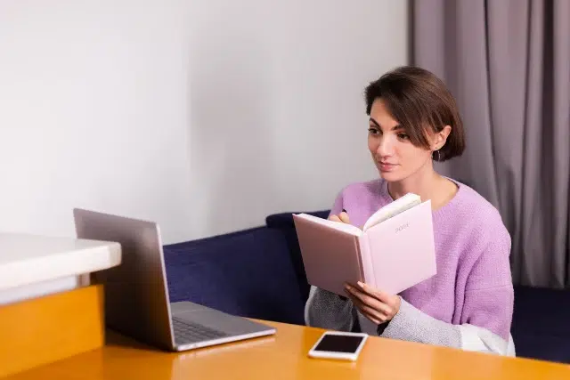 A woman in a lavender sweater sits at a table, attentively reading a pink notebook. Beside her are a silver laptop and smartphone, suggesting a calm, focused work or study session.