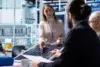 A woman presents a solar panel to three colleagues at a conference table in a modern office. She gestures expressively, suggesting a technical discussion.