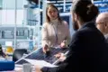 A woman presents a solar panel to three colleagues at a conference table in a modern office. She gestures expressively, suggesting a technical discussion.