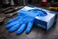 Blue nitrile disposable gloves partially pulled from a Nitri Tech glove box, resting on a dark workbench with hand tools in the background.