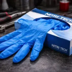Blue nitrile disposable gloves partially pulled from a Nitri Tech glove box, resting on a dark workbench with hand tools in the background.