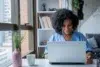 A woman with curly hair and a blue shirt is smiling while working on a laptop at a bright, cozy home office. A plant and mug are on the desk.
