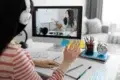 A person wearing headphones waves during an online lesson displayed on a computer screen. A desk with stationery and a clock is visible.