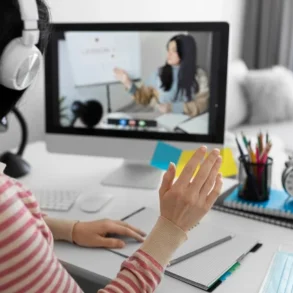 A person wearing headphones waves during an online lesson displayed on a computer screen. A desk with stationery and a clock is visible.