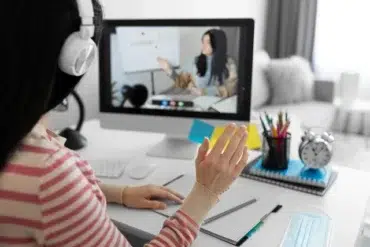 A person wearing headphones waves during an online lesson displayed on a computer screen. A desk with stationery and a clock is visible.