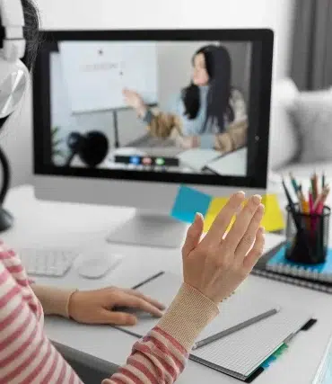 A person wearing headphones waves during an online lesson displayed on a computer screen. A desk with stationery and a clock is visible.