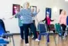 A senior woman with curly white hair and a striped shirt balances on one leg in an exercise class. Surrounding her are other older adults, all participating. The room has a bright and focused atmosphere.