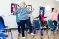 A senior woman with curly white hair and a striped shirt balances on one leg in an exercise class. Surrounding her are other older adults, all participating. The room has a bright and focused atmosphere.