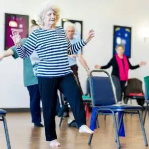 A senior woman with curly white hair and a striped shirt balances on one leg in an exercise class. Surrounding her are other older adults, all participating. The room has a bright and focused atmosphere.
