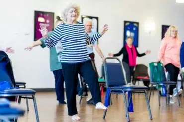 A senior woman with curly white hair and a striped shirt balances on one leg in an exercise class. Surrounding her are other older adults, all participating. The room has a bright and focused atmosphere.