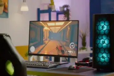 A gaming setup with a glowing desktop PC tower and monitor displaying a first-person shooter game. The room is brightly lit and stylishly decorated.