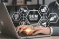 A person types on a laptop with floating hexagonal icons representing SEM concepts, like analytics and advertising. The mood is professional and focused.