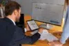 A man in a suit reviews Bitcoin prices on a tablet, pointing to charts. A computer screen shows a rising trend. Papers with graphs are on the table.