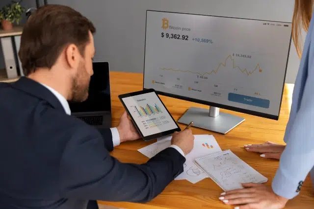A man in a suit reviews Bitcoin prices on a tablet, pointing to charts. A computer screen shows a rising trend. Papers with graphs are on the table.