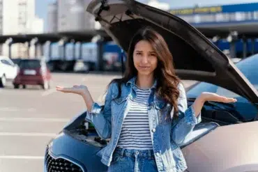 A woman stands confused with outstretched arms in front of a car with an open hood in a parking lot, expressing uncertainty about car trouble.