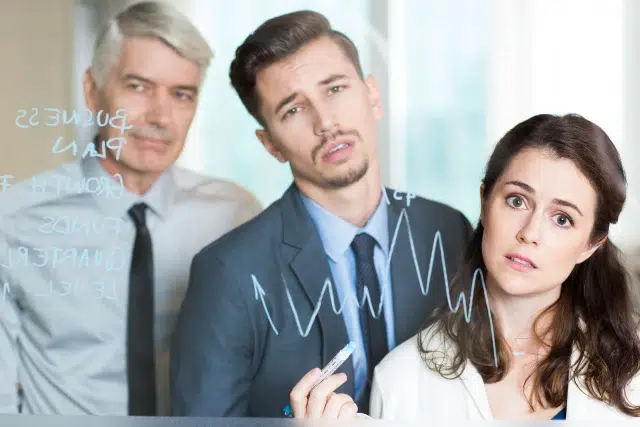 Three people in business attire look concerned while staring at a glass wall with financial graphs and notes. The atmosphere is tense and focused.